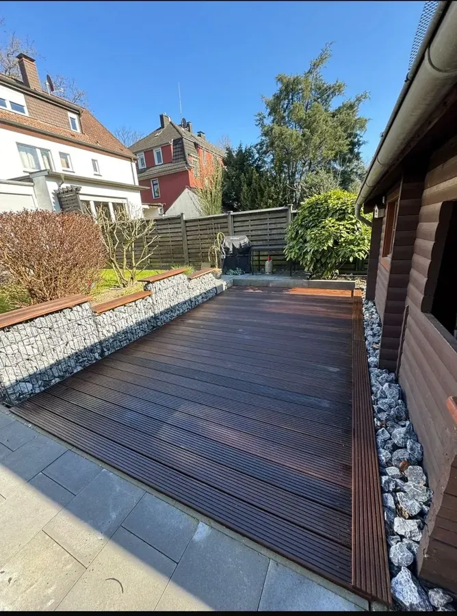 Fertige Terrasse: dunkel geölte Holzdielen, seitlich Gabionenkörbe mit durchgehender Holzauflage als Sitzbank